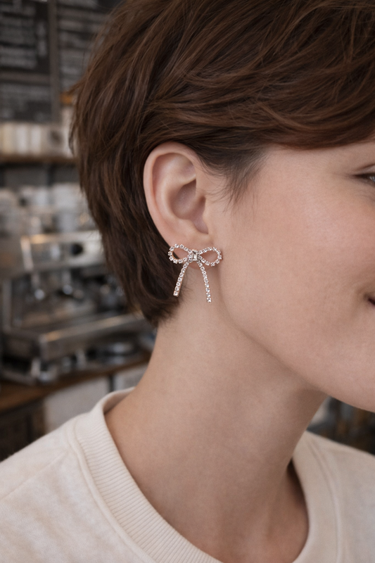 Close-up of a person wearing a bow-shaped rhinestone earring in a blurred indoor setting