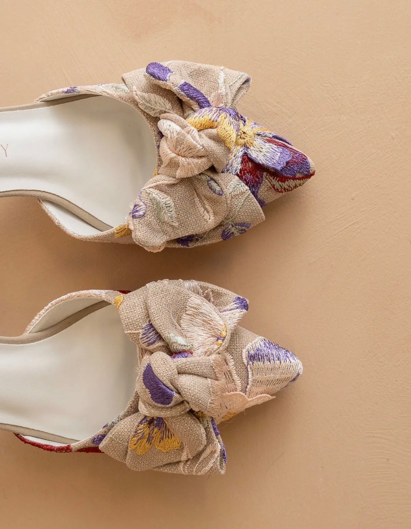 pair of high-heeled slip on shoes with floral tapestry fabric with bow accent on a beige background