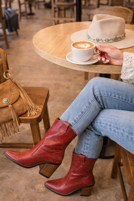 Person wearing red cowboy snake skin embossed ankle boots sitting at a cafe table with a cup of coffee.