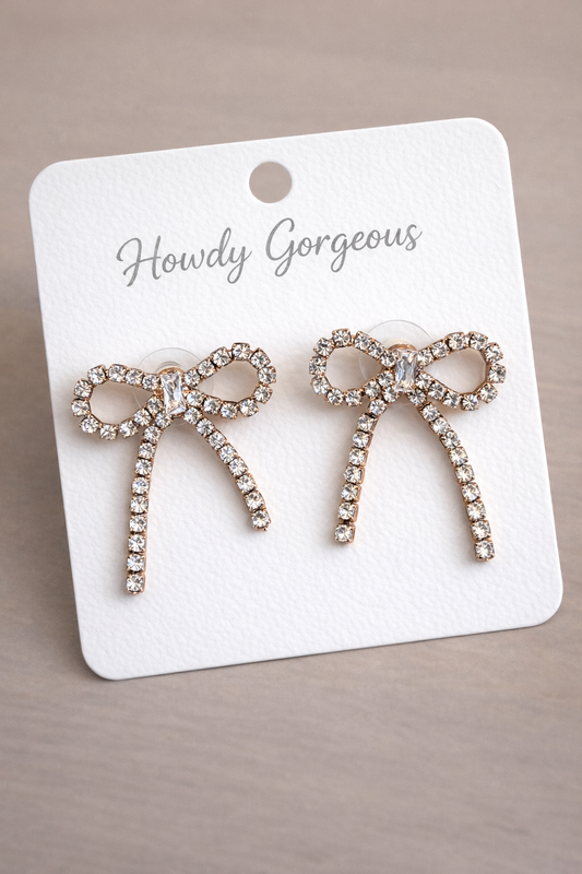 Bow-shaped rhinestone stud earrings on a branded card against a beige background